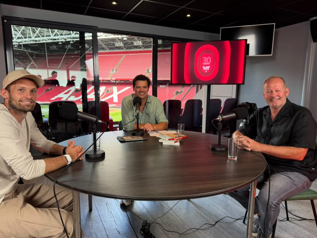 Drie mannen aan tafel met microfoons tijdens een podcastopname in een studio met uitzicht op het stadion