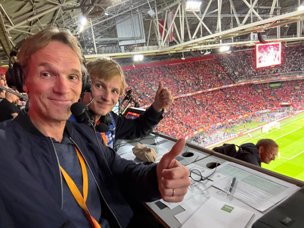 Twee audiodescriptie-commentatoren met headset op de perstribune van een vol voetbalstadion, duim omhoog