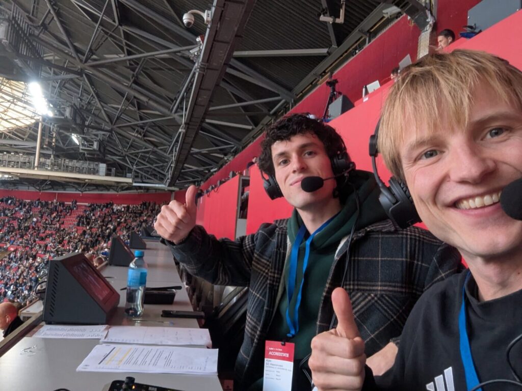 Daaf en Diederik met headset op de perstribune van het stadion, duim omhoog